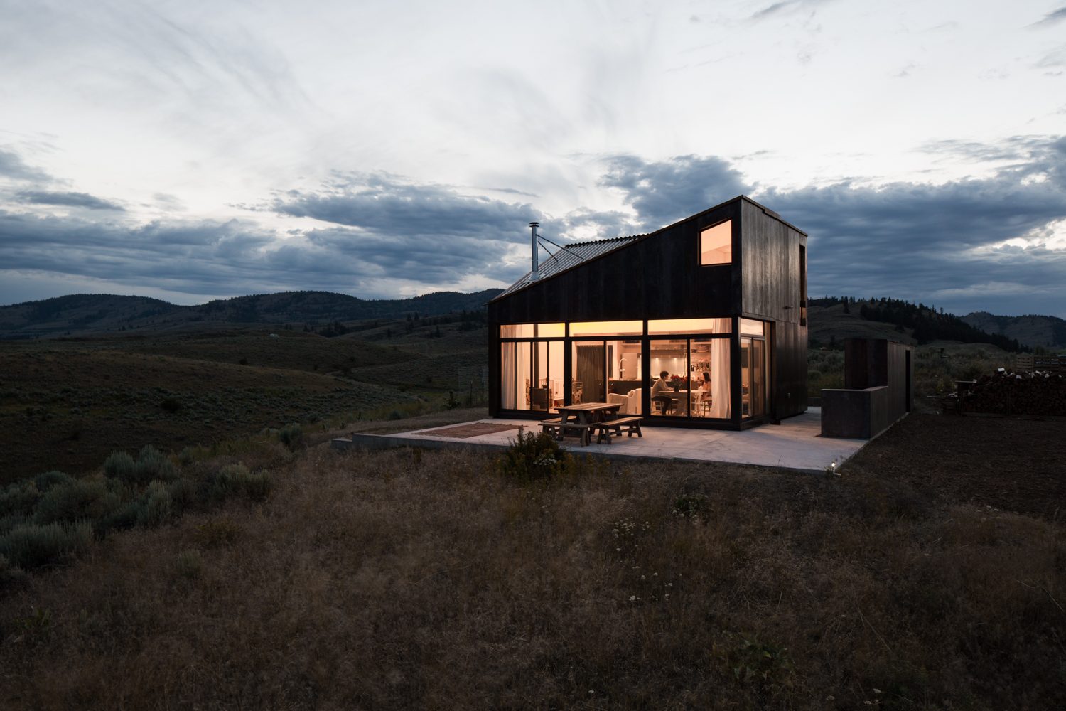 Sky House at dusk, its glass wall lit from within against the hills of the Okanogan Highlands